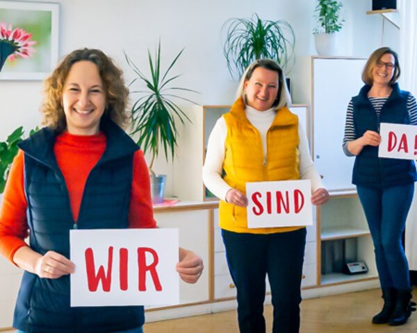 Drei freundliche Frauen halten Schilder mit den Worten "Wir sind da" hoch | © Stefan Dehmel