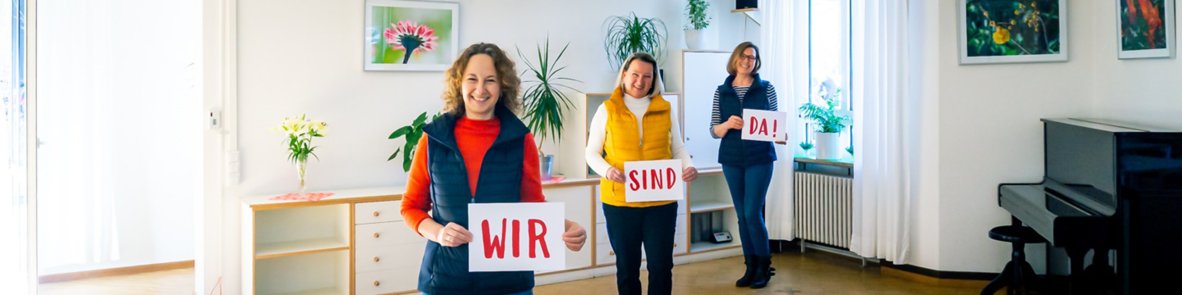 Drei freundliche Frauen halten Schilder mit den Worten "Wir sind da" hoch | © Stefan Dehmel